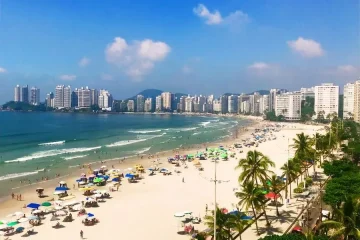 Vista aérea da praia do Guarujá, SP. Orla com prédios, mar azul, areia clara, guarda-sóis e palmeiras. Turismo no litoral paulista.
