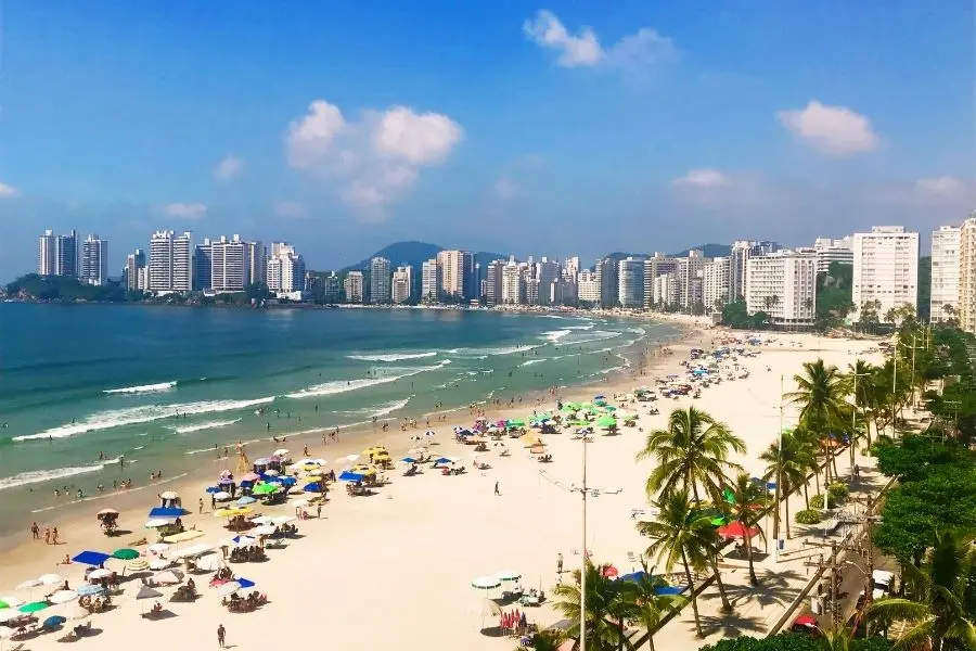 Vista aérea da praia do Guarujá, SP. Orla com prédios, mar azul, areia clara, guarda-sóis e palmeiras. Turismo no litoral paulista.