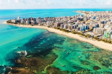 Vista aérea da Praia do Francês, Alagoas. Mar azul-turquesa, recifes, barcos e prédios na orla. Paraíso tropical!