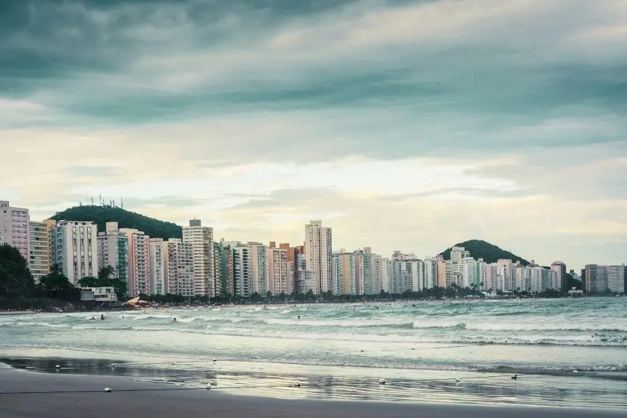 Vista da Praia do Tombo, Guarujá. Prédios, mar com ondas, céu nublado. Paraíso para surfistas e turistas.